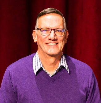 PAH patient, Steve, wearing glasses and a purple sweater while smiling in front of red curtains on a theatre stage