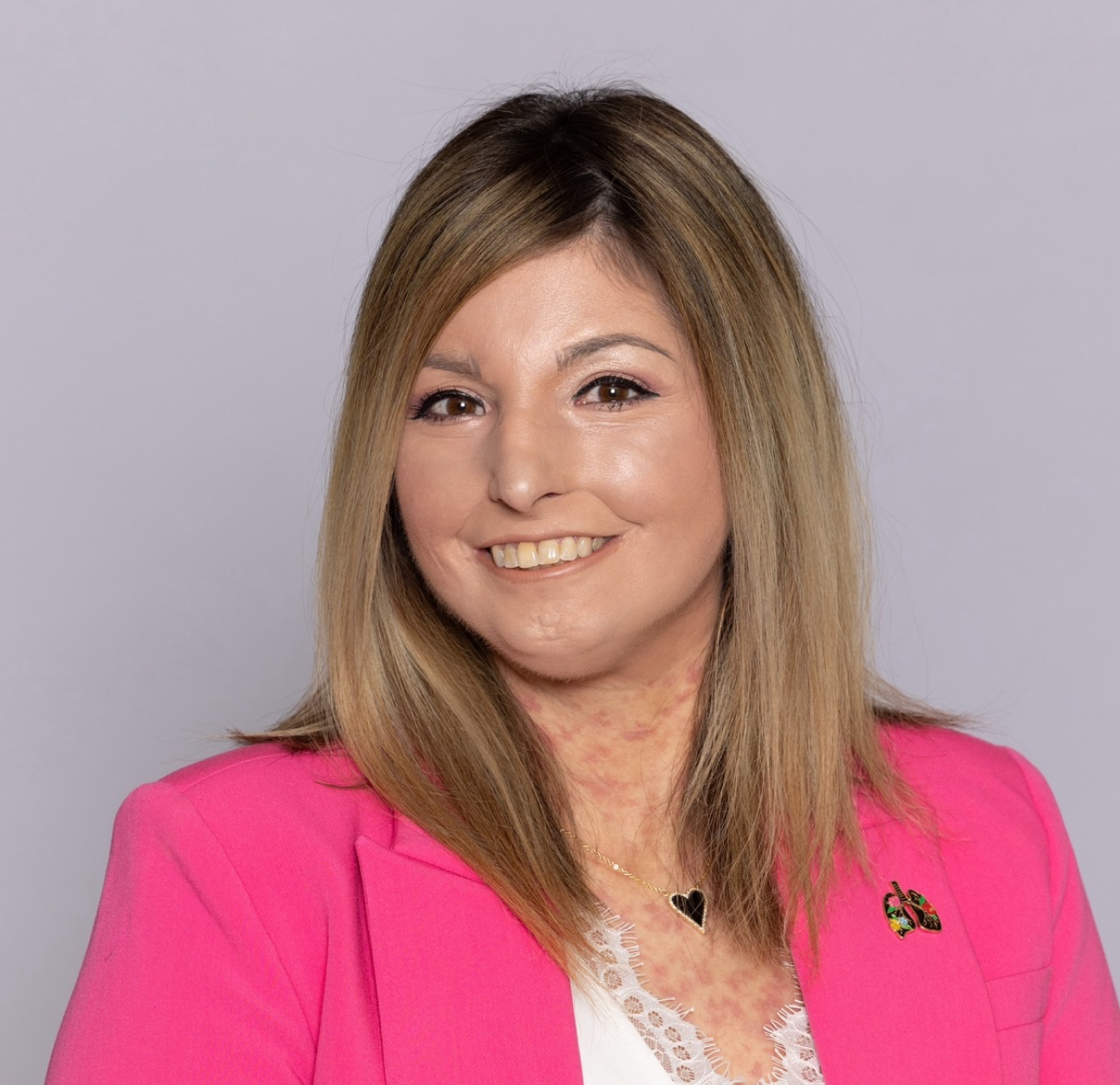 Scleroderma and PAH patient, Amy, smiling warmly in front of a grey backdrop wearing a bright pink blazer and white shirt