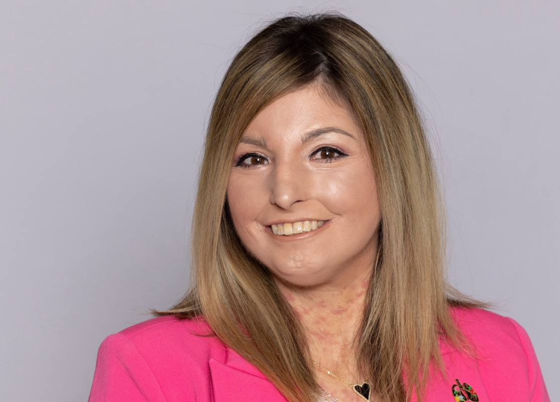 Scleroderma and PAH patient, Amy, smiling in front of a grey backdrop wearing a bright pink blazer and white shirt