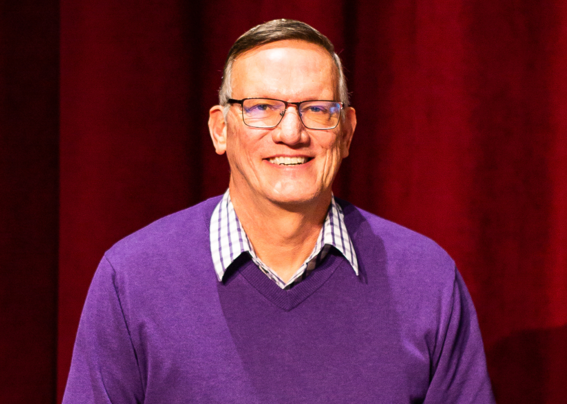 PAH patient, Steve, wearing glasses and a purple sweater while smiling in front of red curtains on a theatre stage