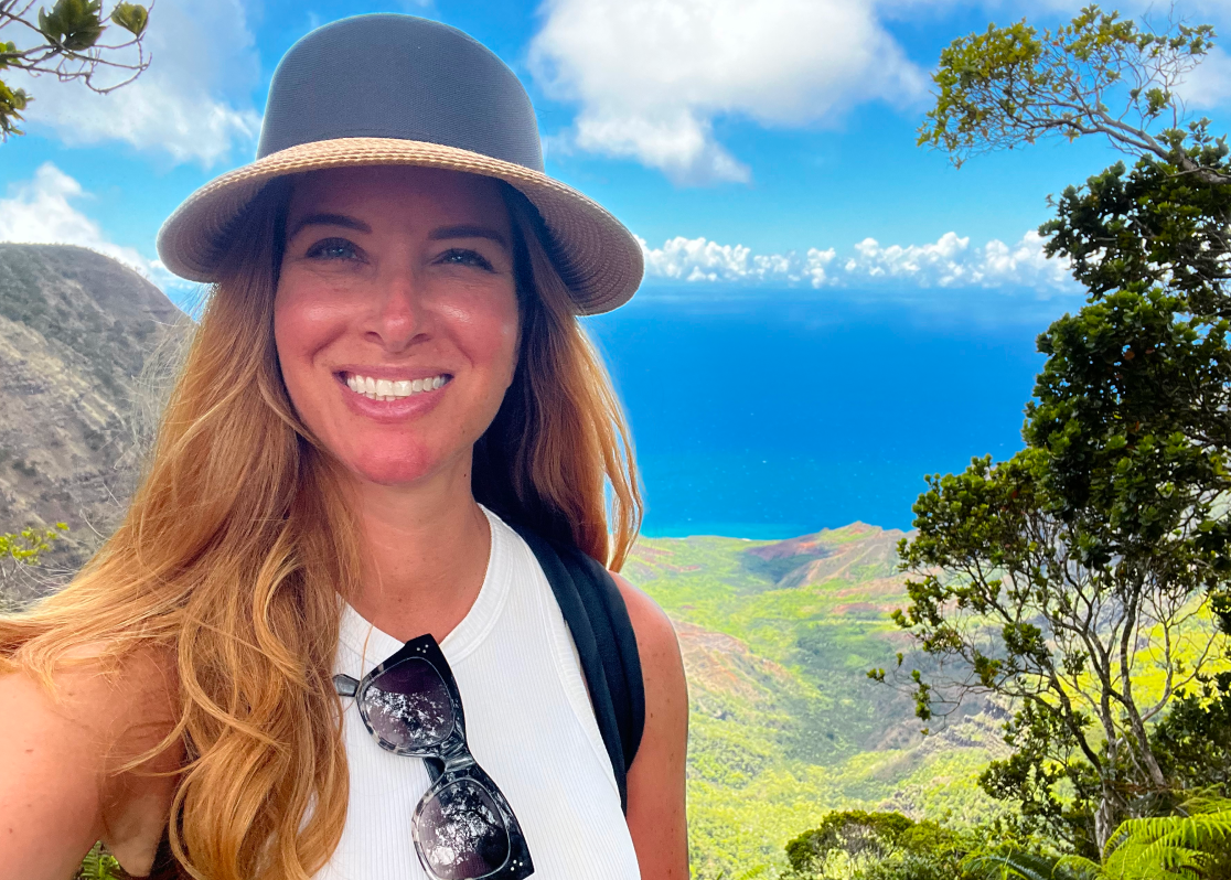 PAH patient, Katie, smiling while hiking in the mountains on a sunny day wearing a straw hat and backpack