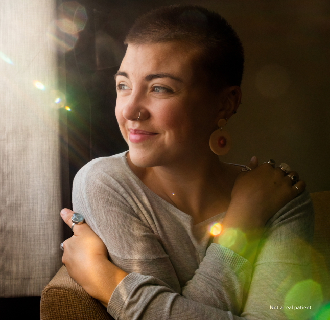 A woman with short hair smiling, glancing to her right with arms wrapped around herself while sitting in the sunlight