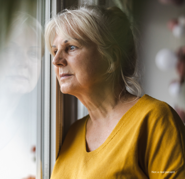 An elderly woman wearing a yellow shirt gazing out the window with a calm expression and her hands folded together