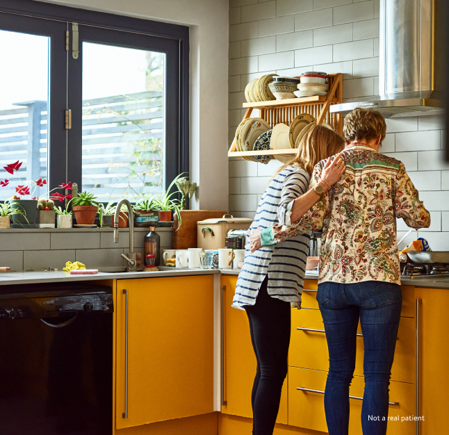 Two women side-by-side looking into a pot at the stove with both of their hands touching each others backs