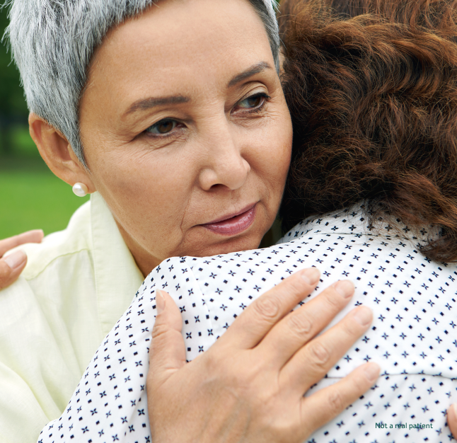 A grey haired woman facing the camera, tightly hugging another woman with brown hair outside