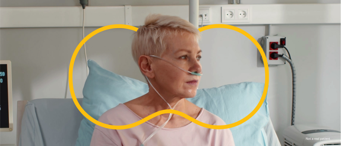 A female patient wearing oxygen tubes sitting on a hospital bed with the yellow Outnumber PAH symbol around her