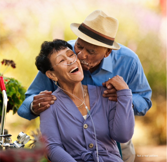 An elderly man, wearing a hat, kisses an elderly woman on the cheek who is smiling and wearing oxygen tubes while outdoors