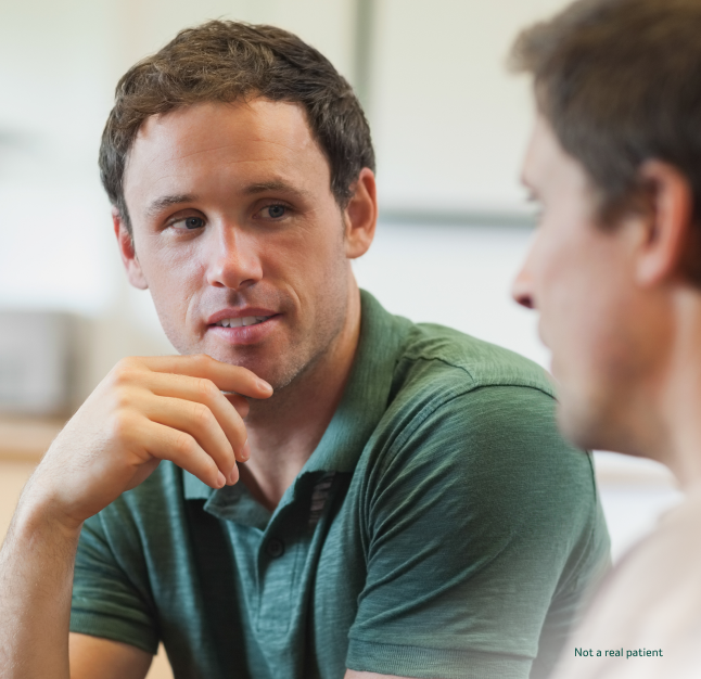 A man wearing a green polo shirt rests his hand on his chin while listening thoughtfully to a another man talking