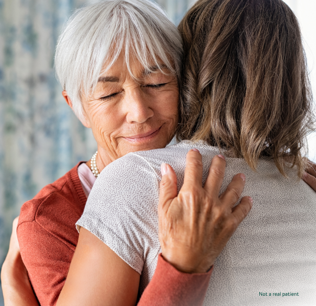 A smiling older woman with grey hair hugging another woman with eyes closed