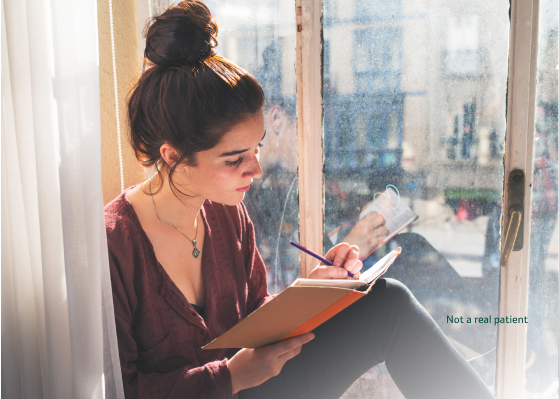 A young woman wearing a red sweater sitting on a windowsill while writing in a notebook