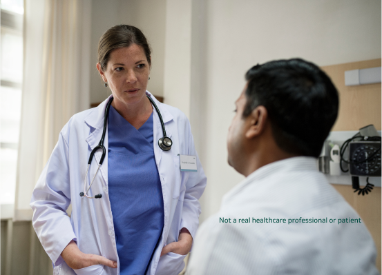 A female doctor in blue scrubs and a white lab coat wearing a stethoscope around her neck is talking to a male patient