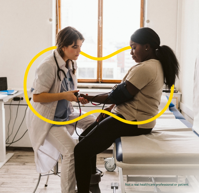 Female doctor checks blood pressure of female patient sitting on exam room table with yellow Outnumber PAH logo around them