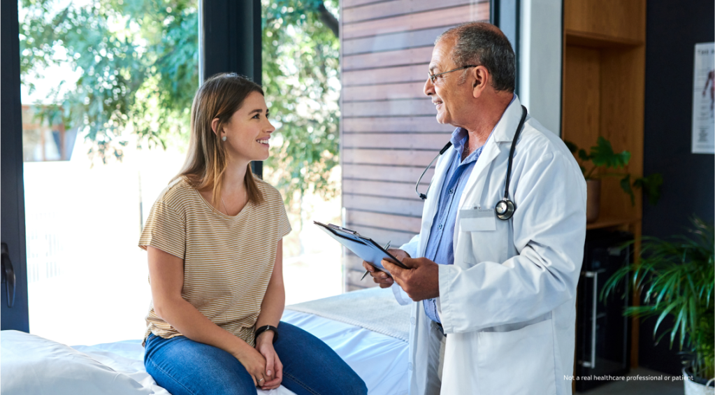 A female patient sitting and smiling on an examination table while consulting with a male doctor in front of a sunny window